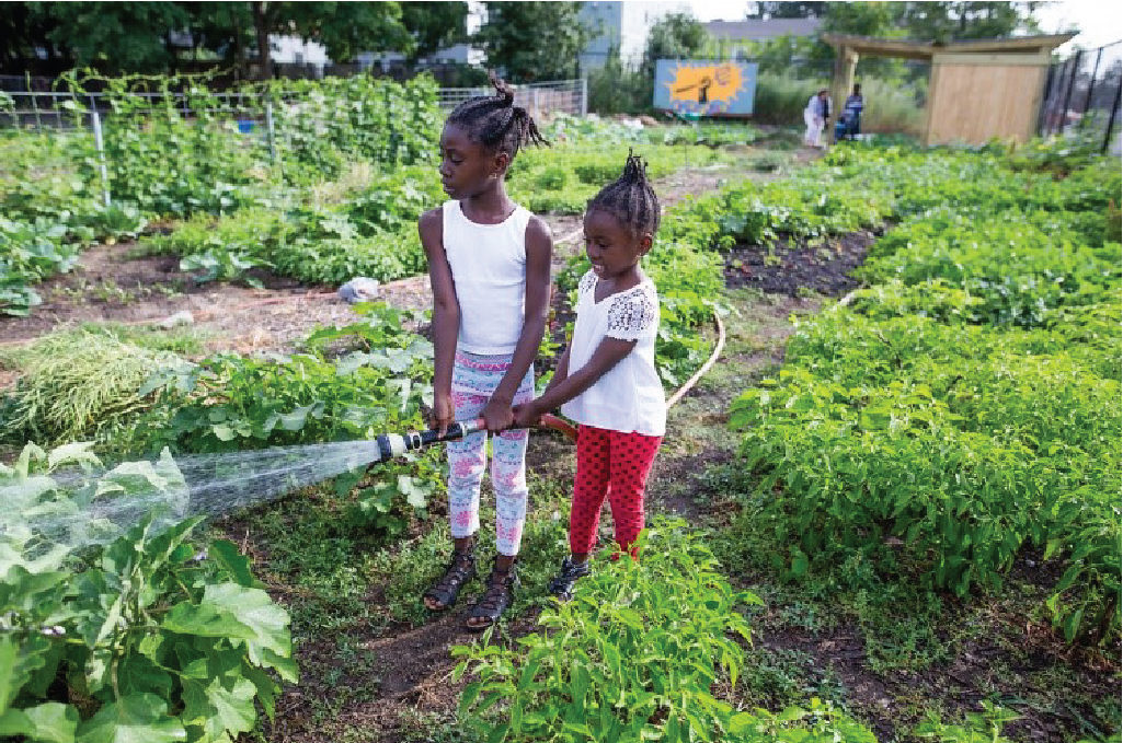 Two young girls water a community garden with a host