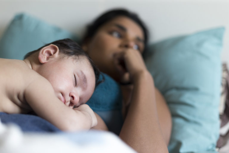 Baby asleep in bed on his mother's chest, who is enjoying the break from stressful parenting.