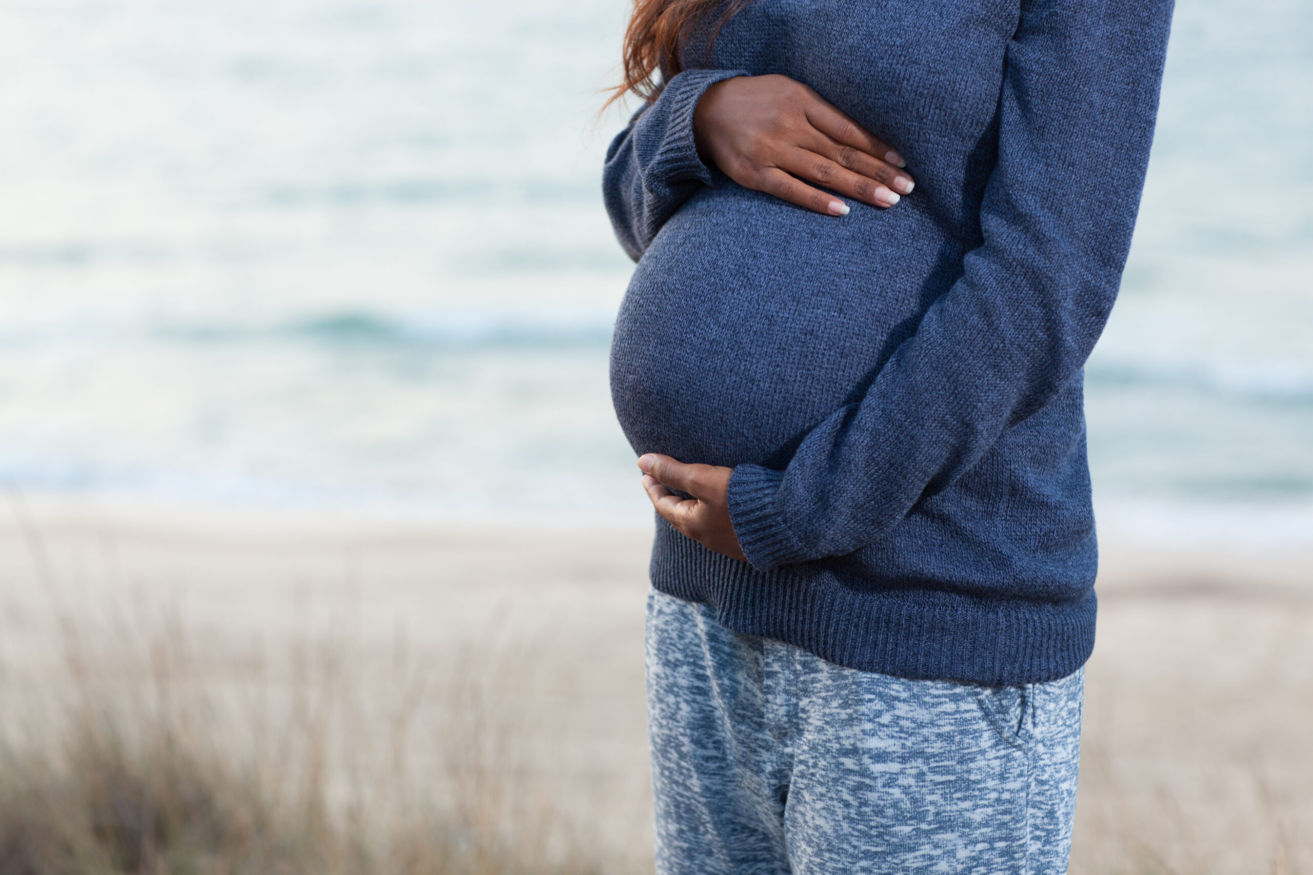 Close up on pregnant woman belly. Unrecognized pregnant woman in her trimester touching and stroking her big belly, standing on a sea shore.
