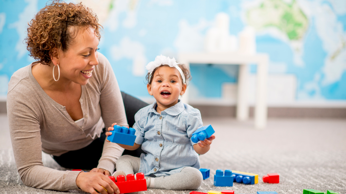 Child sitting on the ground smiling and holding building blocks beside woman.