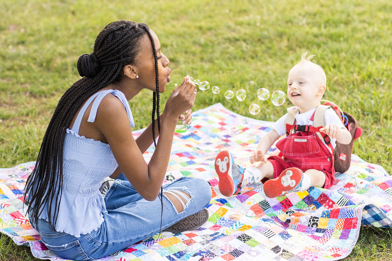 young adult in blue top blows bubbles next to smiling child in red overalls on patch blanket in grass field