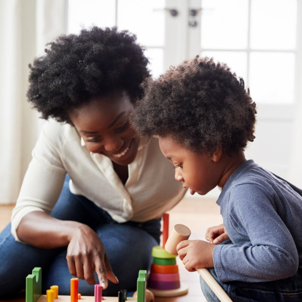 A black mother with an Afro sits on the ground while playing a game with blocks with her child. Her child also has an Afro and squats while holding a mallet in their hand for the game. 