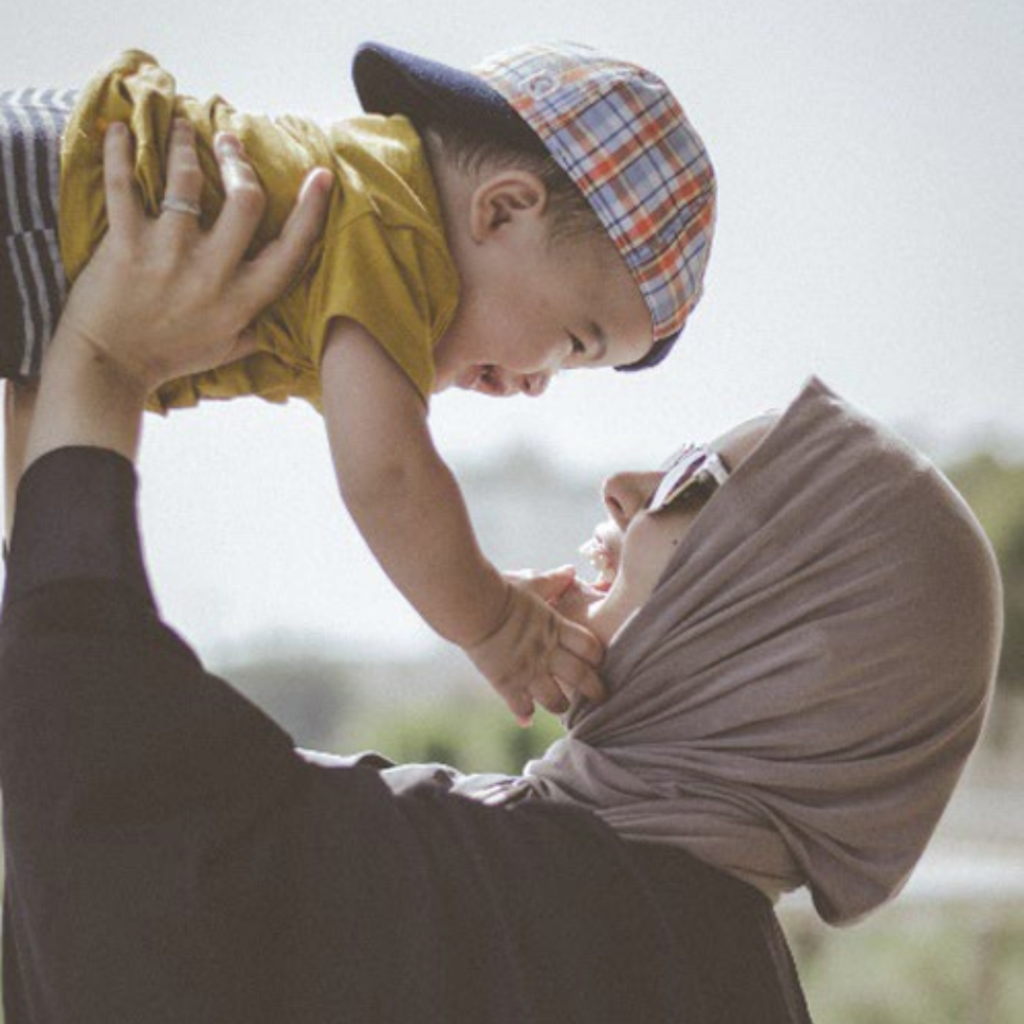A Muslim woman wearing a hijab and sunglasses lifts her baby above her head. The baby is wearing a backwards baseball cap and touching their mother's chin with their hands while they laugh and smile.