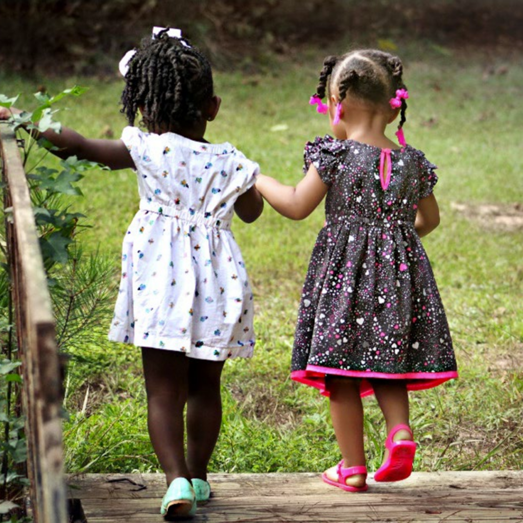 Two young girls with dark and light brown skin are wearing dresses and holding hands while crossing a wooden bridge. The girls are walking away from the camera.
