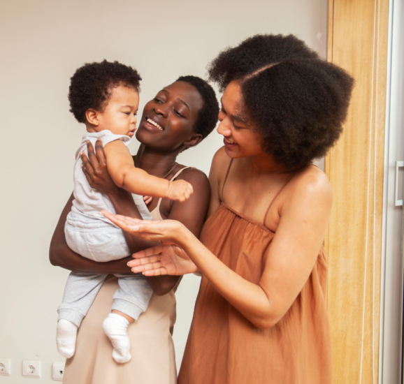 Photo image of a black woman smiling and holding a baby while another black woman holds out her hands to the baby.