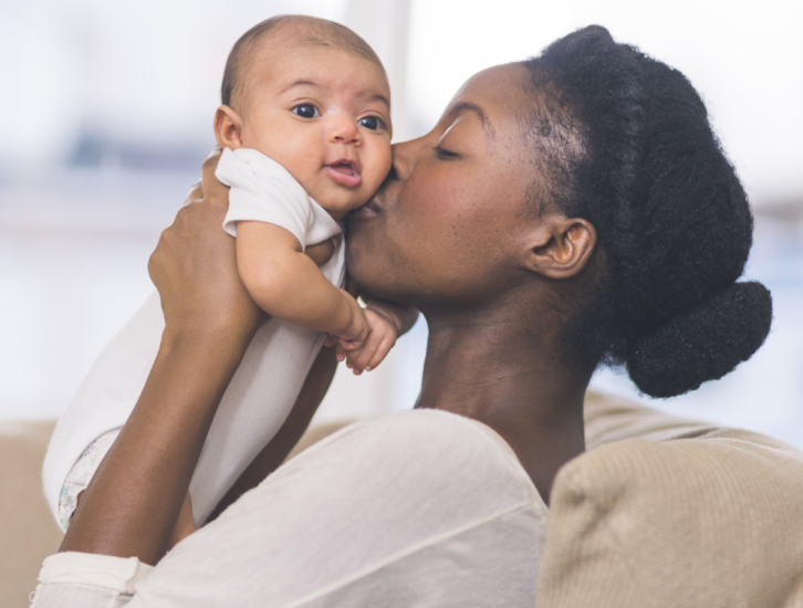 Photo image of a black woman kissing her baby on the cheek. 