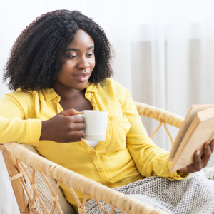 Portrait of smiling woman reading a book, drinking hot coffee, holding cup, and sitting in wicker chair with a blanket.